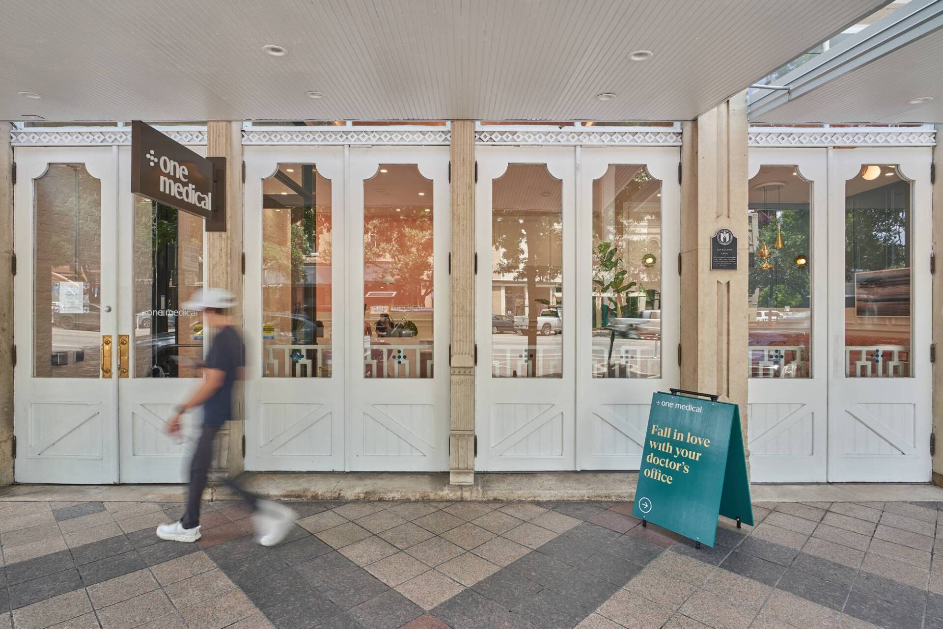 Exterior of a cafe with white doors and glass panels. A person walks in front. Green A-frame sign.