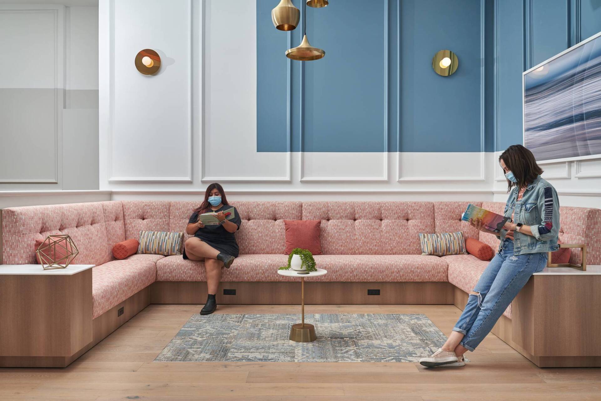 Two people wearing masks read on a pink couch in a lounge with blue and white walls.