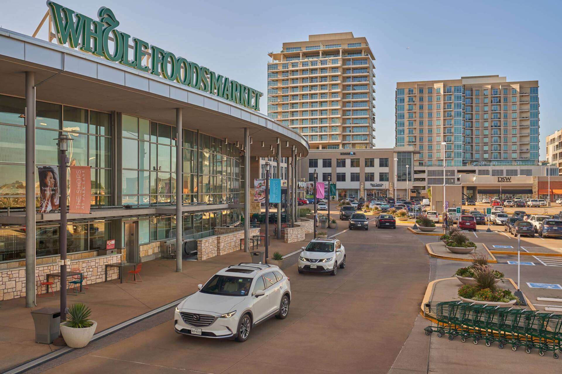 Whole Foods Market exterior with cars driving in front. Tall buildings in the background. Sunny day.