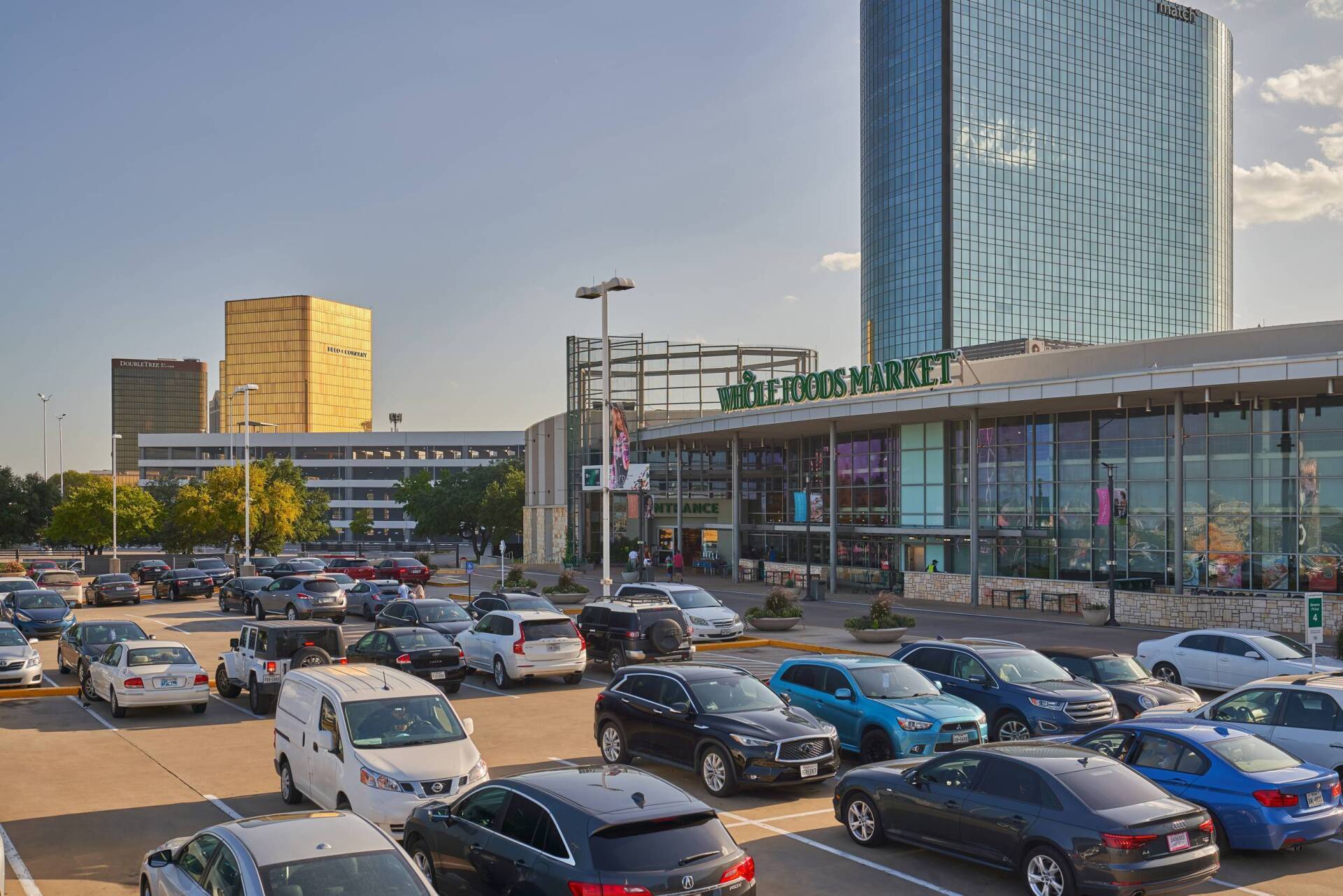 Cars parked in front of Whole Foods Market, with a tall glass skyscraper and golden building in the background.