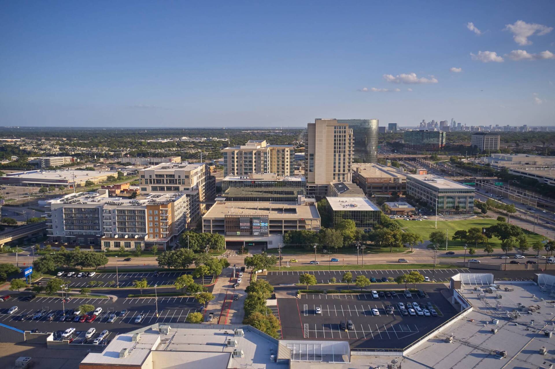 Aerial view of a city with buildings, parking lots, trees, and highway under a clear, blue sky.