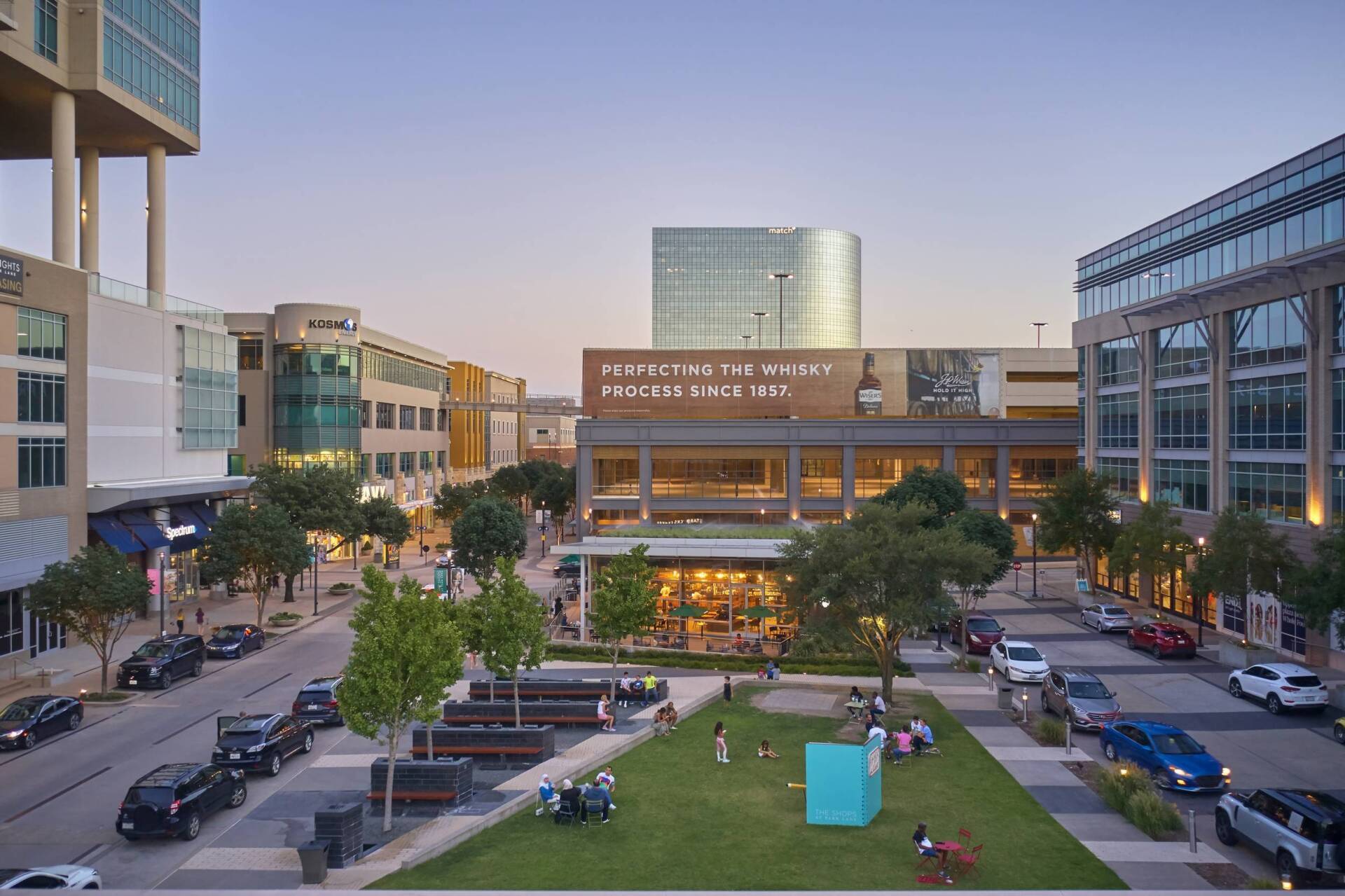 Downtown square with buildings, trees, parked cars, and people on grass at dusk.