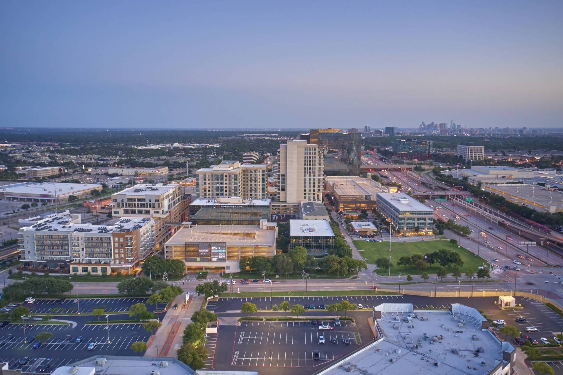 Aerial view of urban area with buildings, parking, and a distant skyline under a dusk sky.