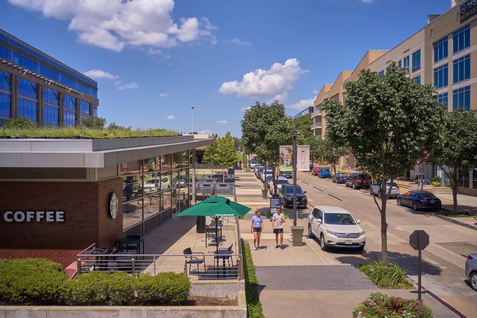 Starbucks with green roof and patio, street with cars, and two people walking on a sunny day.