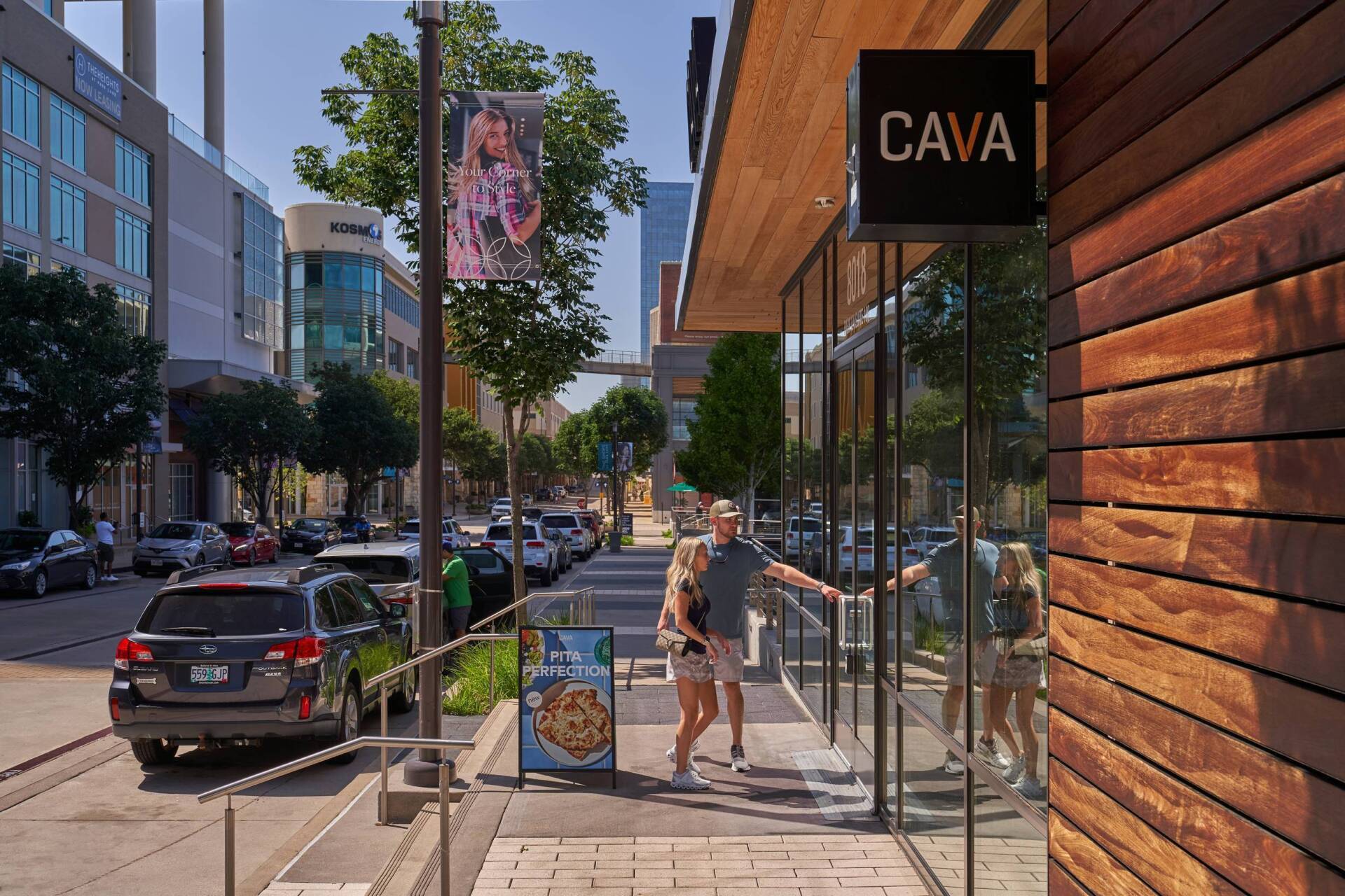 A person opens the door of CAVA restaurant on a sunny street, with cars and buildings.