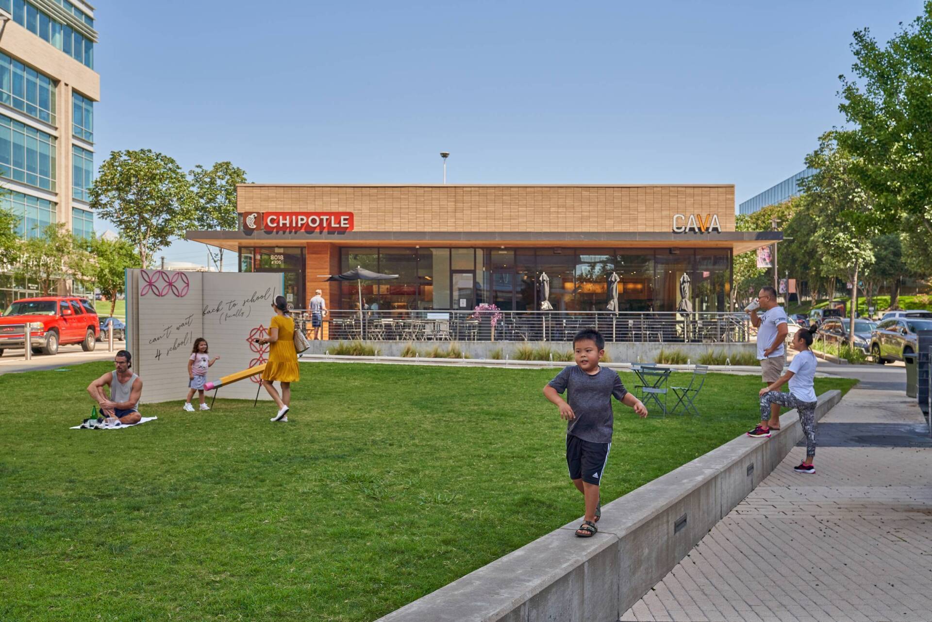 People in a park with a Chipotle restaurant in the background. Children play while others sit on the grass.