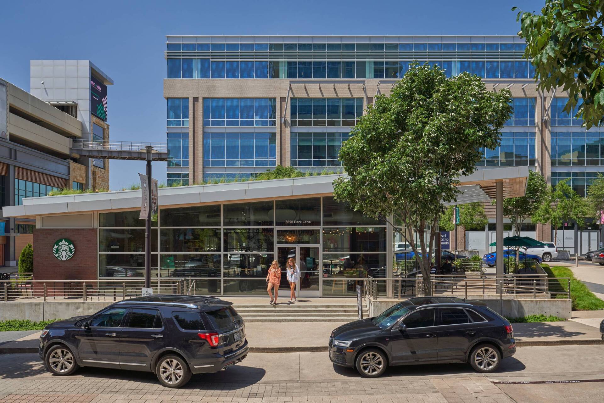 Starbucks storefront with two people entering, flanked by parked cars, in front of a modern building on a sunny day.