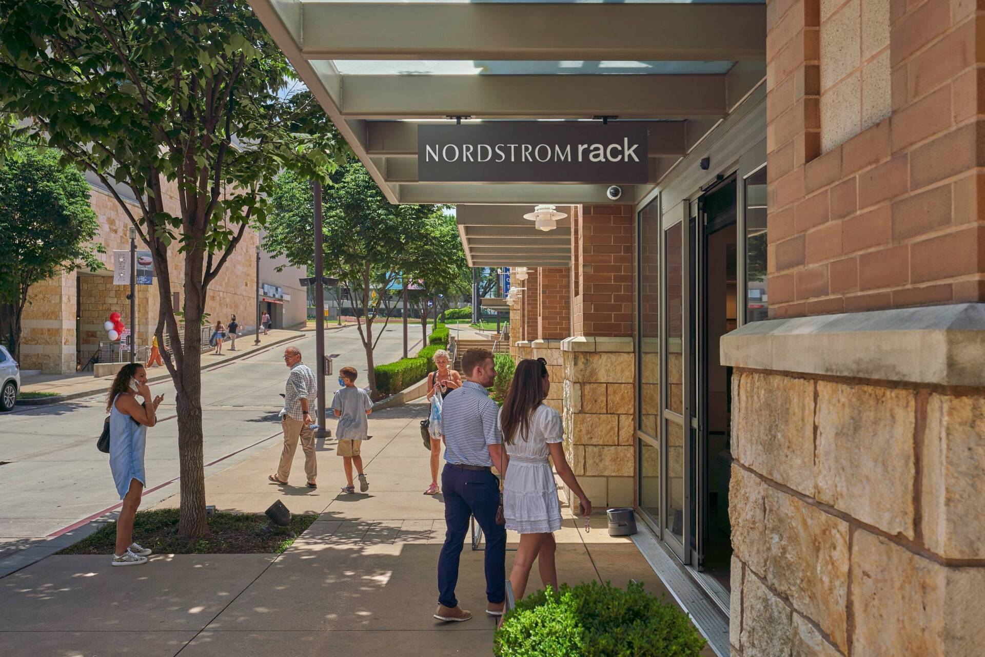 Nordstrom Rack store entrance, people walking in. Exterior brick, sidewalk, trees.