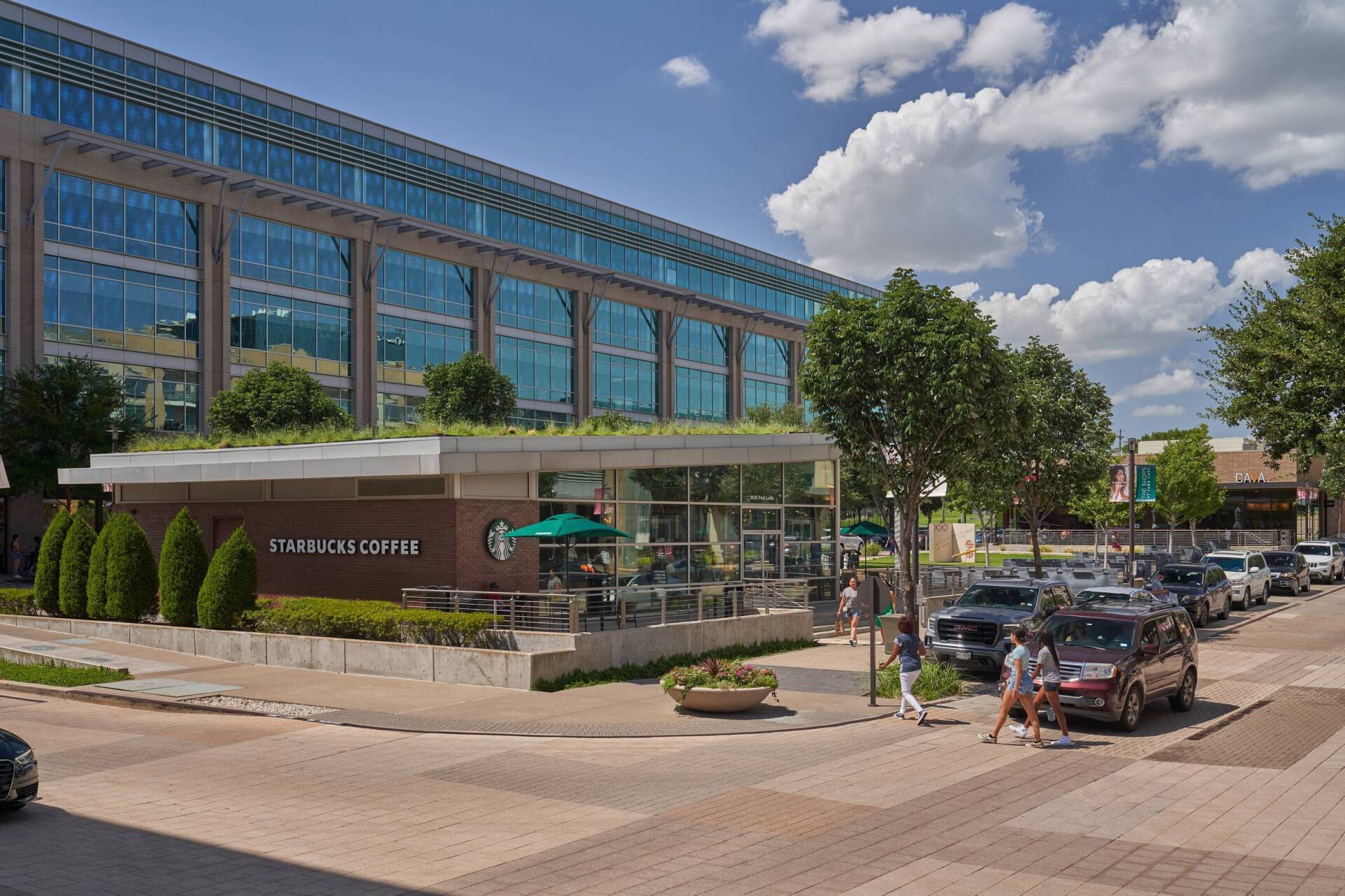 Starbucks cafe with green roof in front of a modern building on a sunny day. People walk on sidewalk. Cars parked nearby.