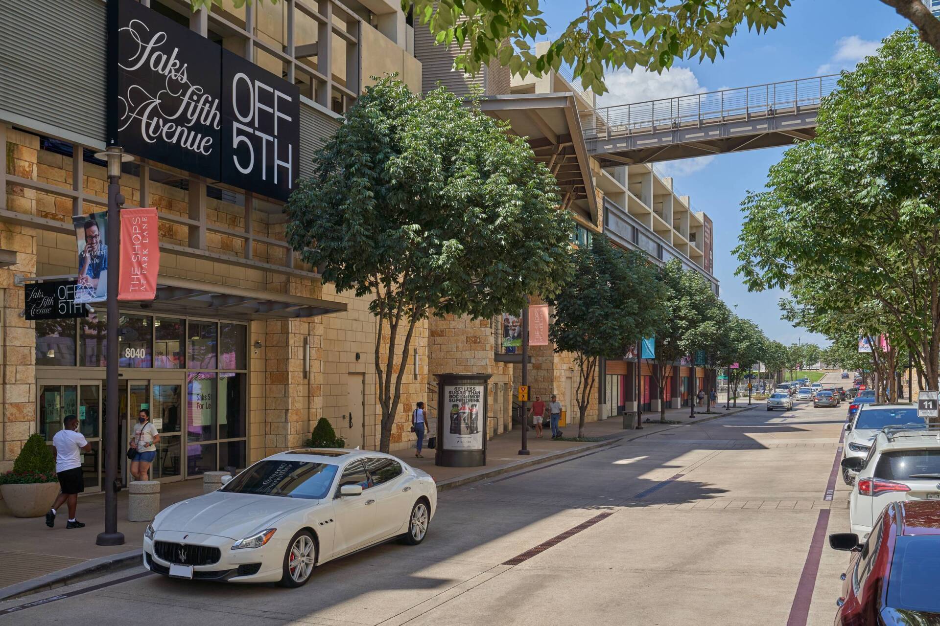 A street with a Saks OFF 5TH store, parked cars, trees, and people. A white Maserati is parked out front.