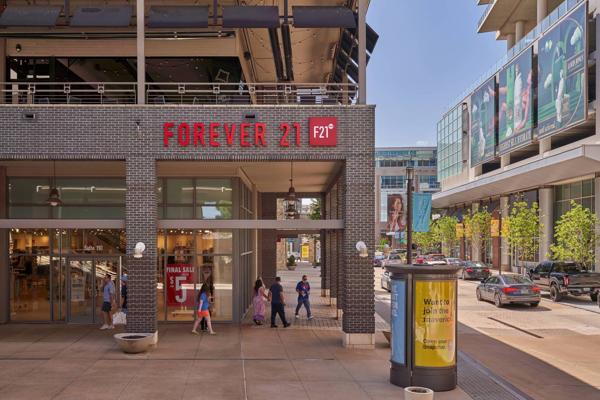 Forever 21 storefront with people walking. Exterior shot on a sunny day. Buildings and street are in view.