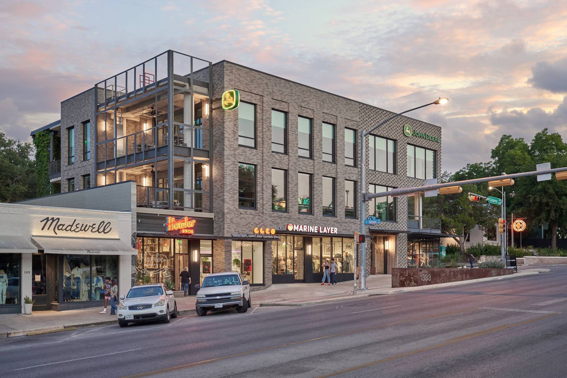 Street view of a three-story brick building with storefronts. Cars parked on the street. Cloudy sunset.