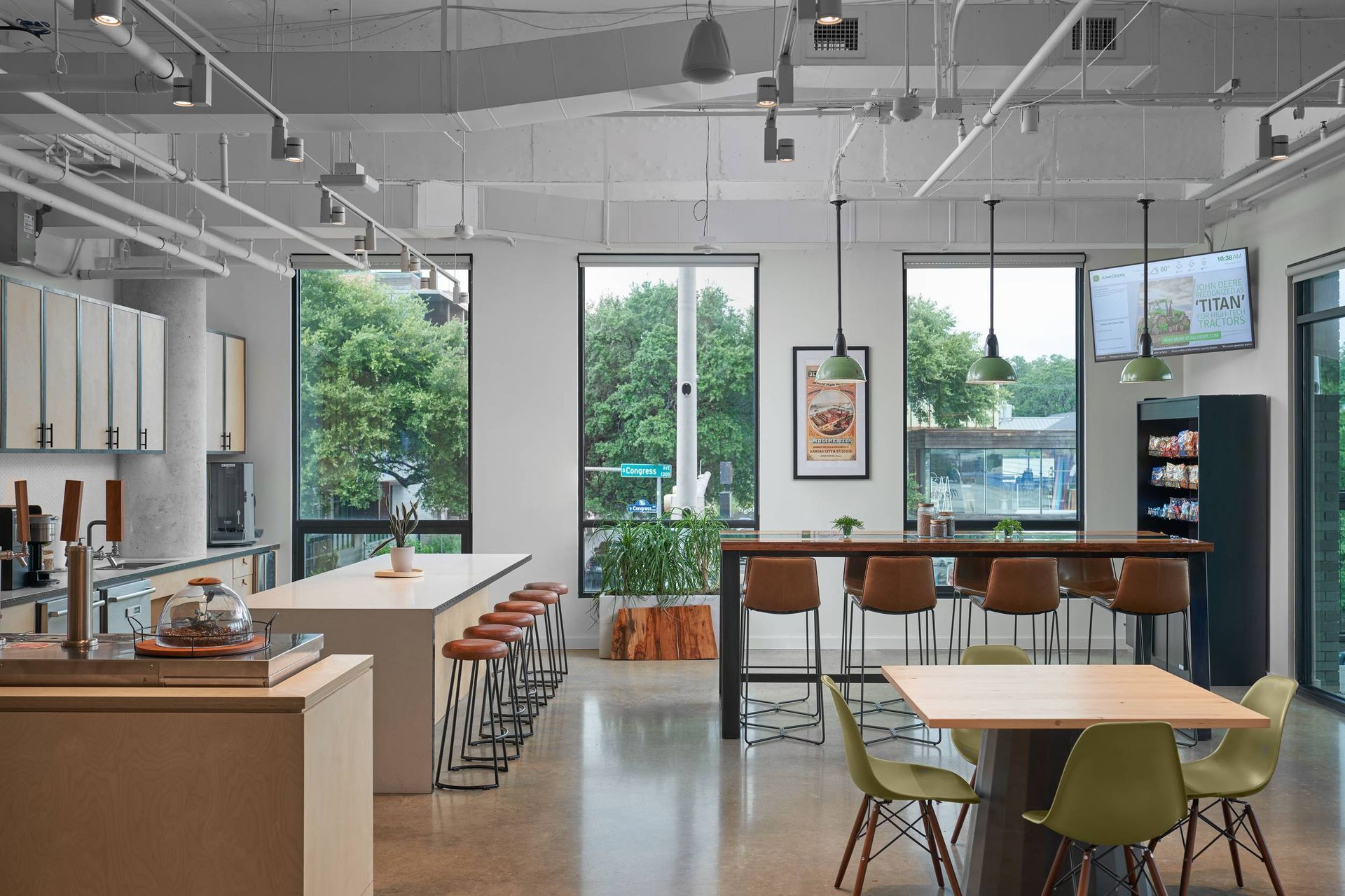 Modern office break room with tables, chairs, bar stools, and windows overlooking greenery.