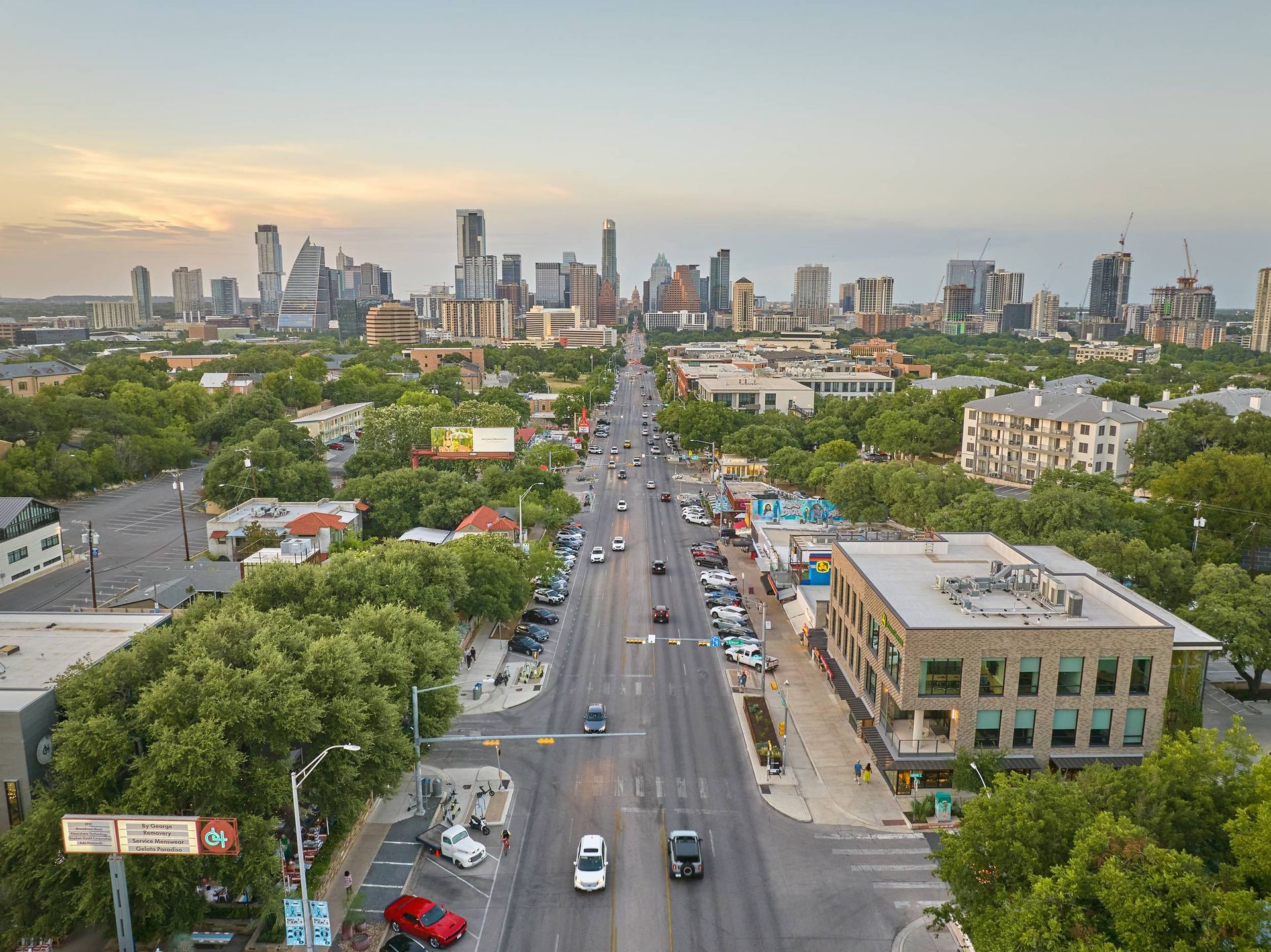 Aerial view of a city street lined with trees, leading to a downtown skyline under a soft sky.
