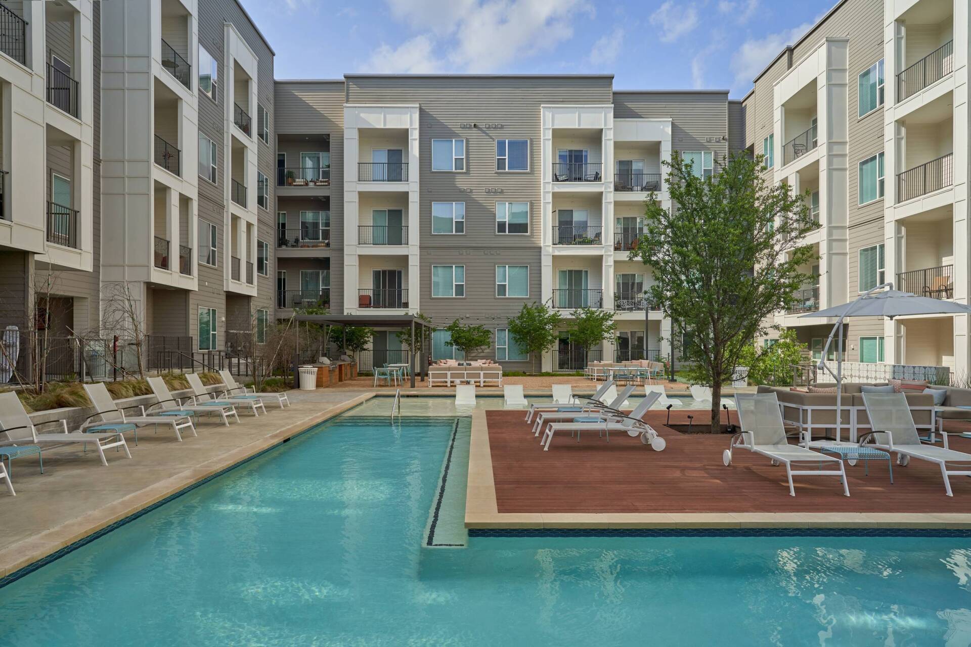 Pool area with lounge chairs, surrounded by modern apartment buildings and a tree.