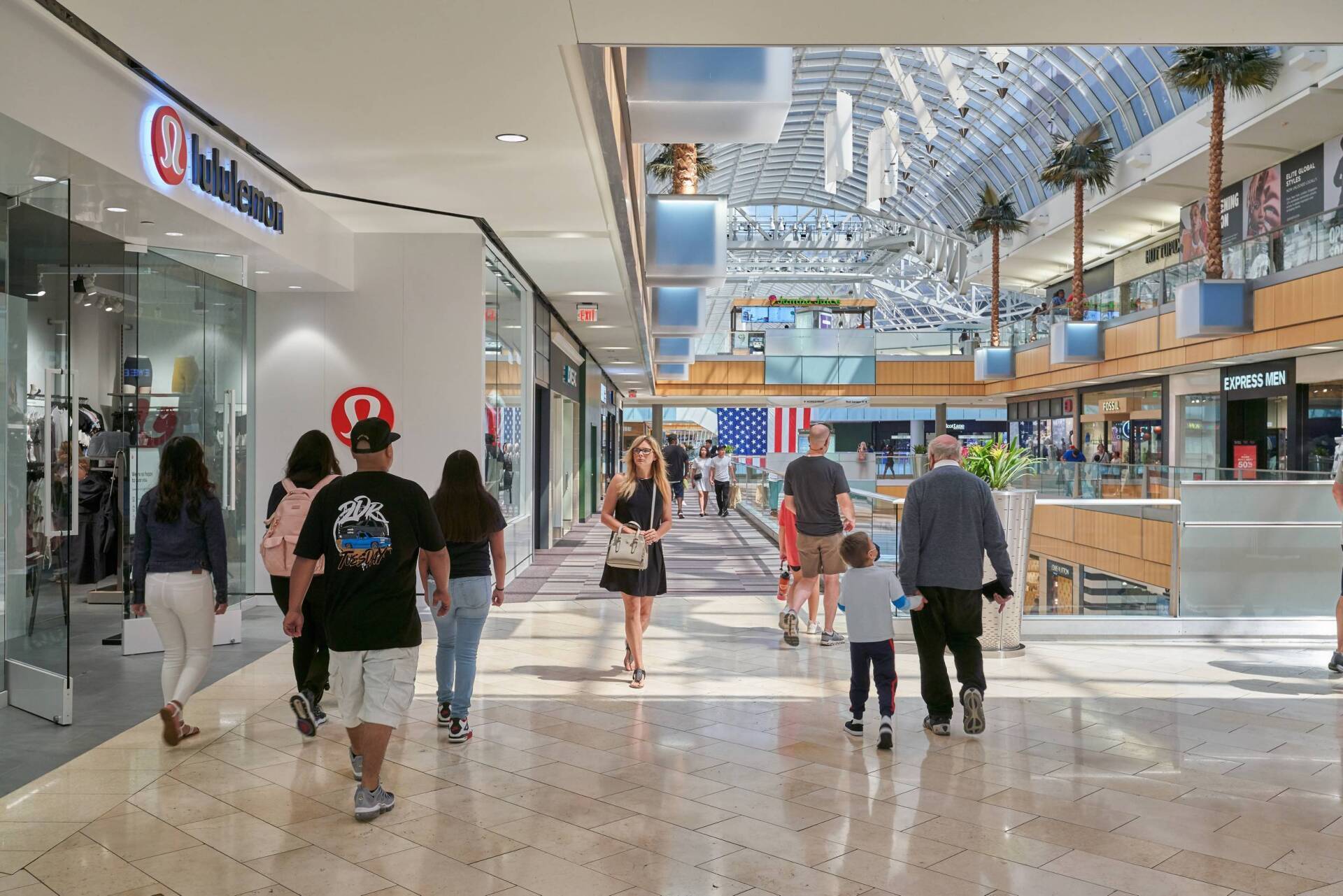 People walking through a brightly lit shopping mall with various store fronts.