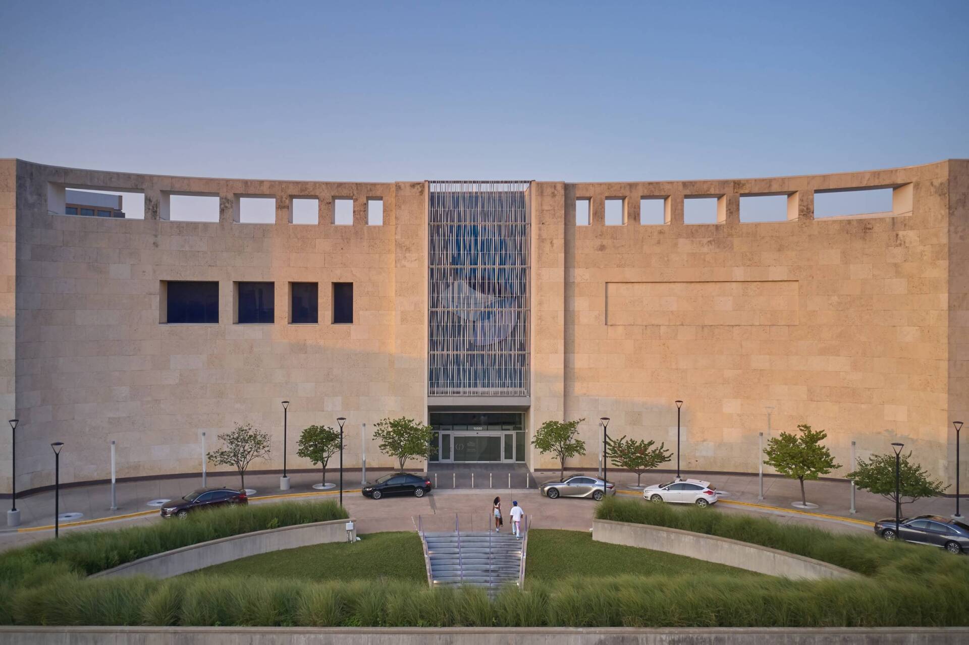 Stone building with central glass entrance, curved facade, small windows, and landscaped area.