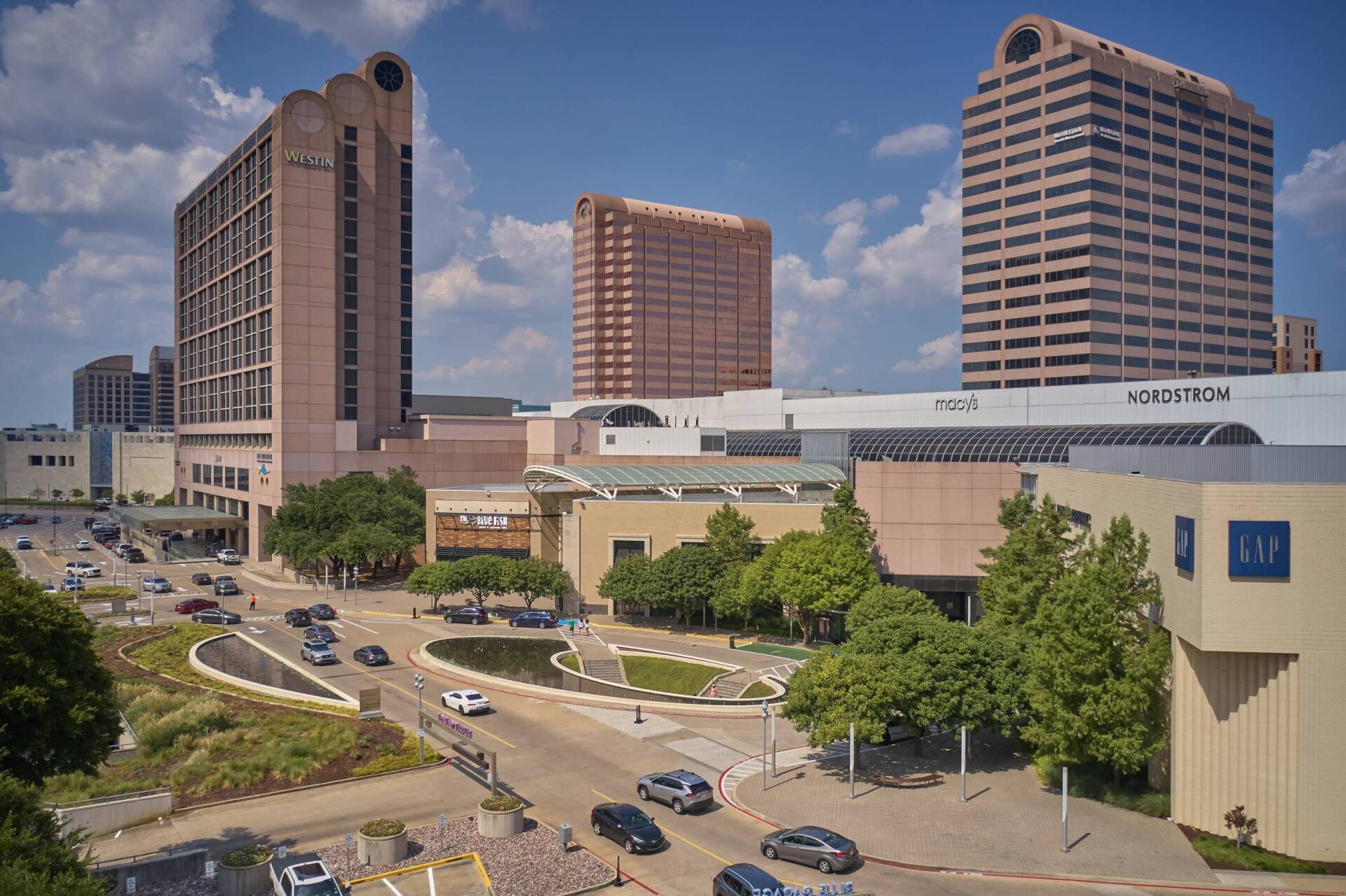 Cityscape with buildings of varying heights under a blue sky, including a hotel and a mall. Cars on the road.