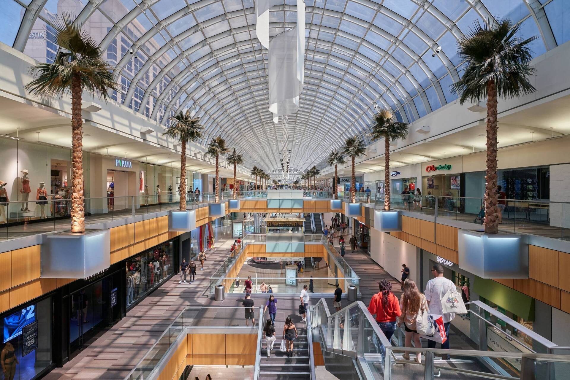 Interior view of a large shopping mall with multiple levels, glass ceiling, palm trees, and people walking.