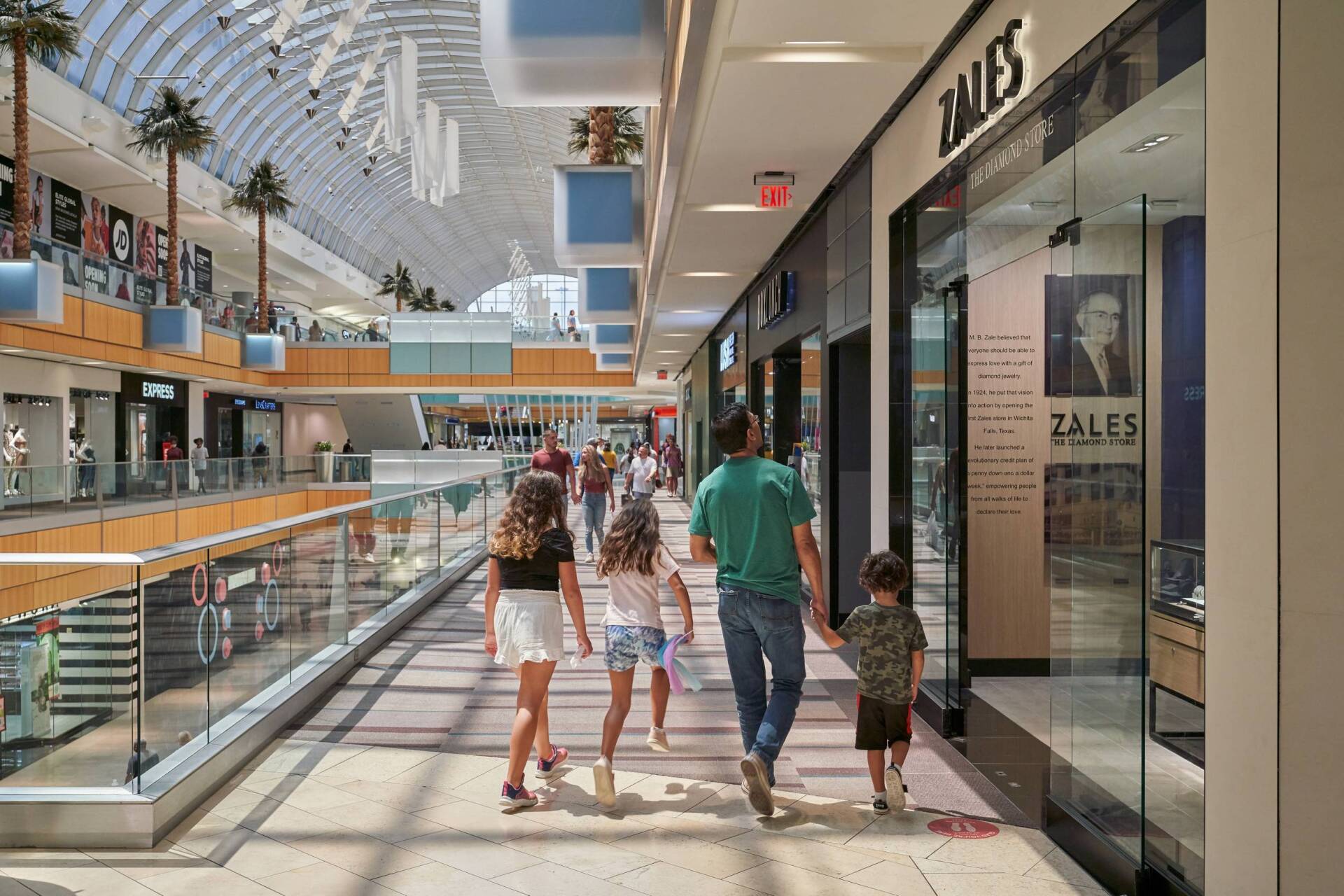 Family walking in a mall, passing Zales jewelry store. Bright interior, glass railings, palm trees, arched ceiling.