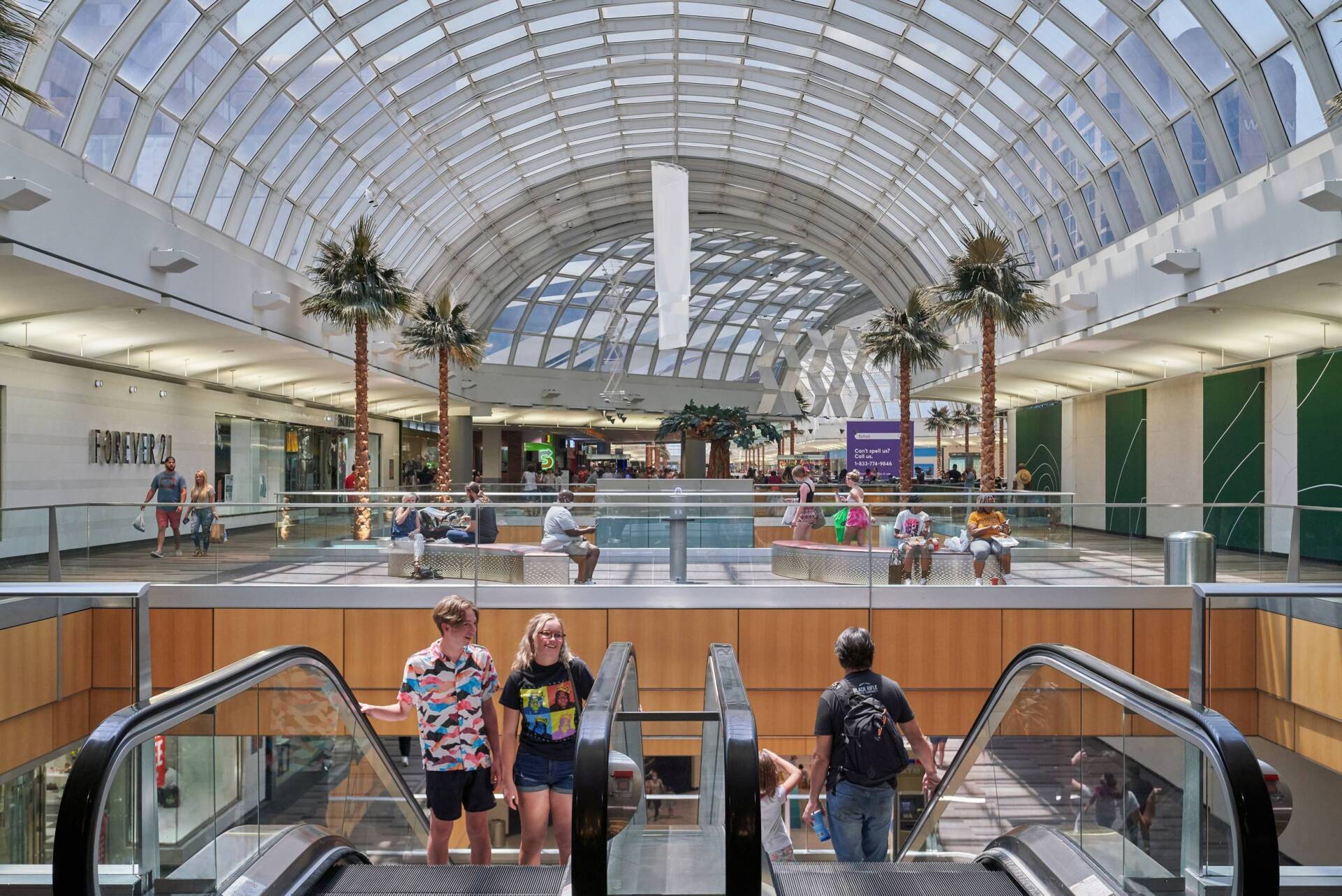 Interior of a mall with an arched glass roof. People ride escalators. Palm trees and shops line the floors.