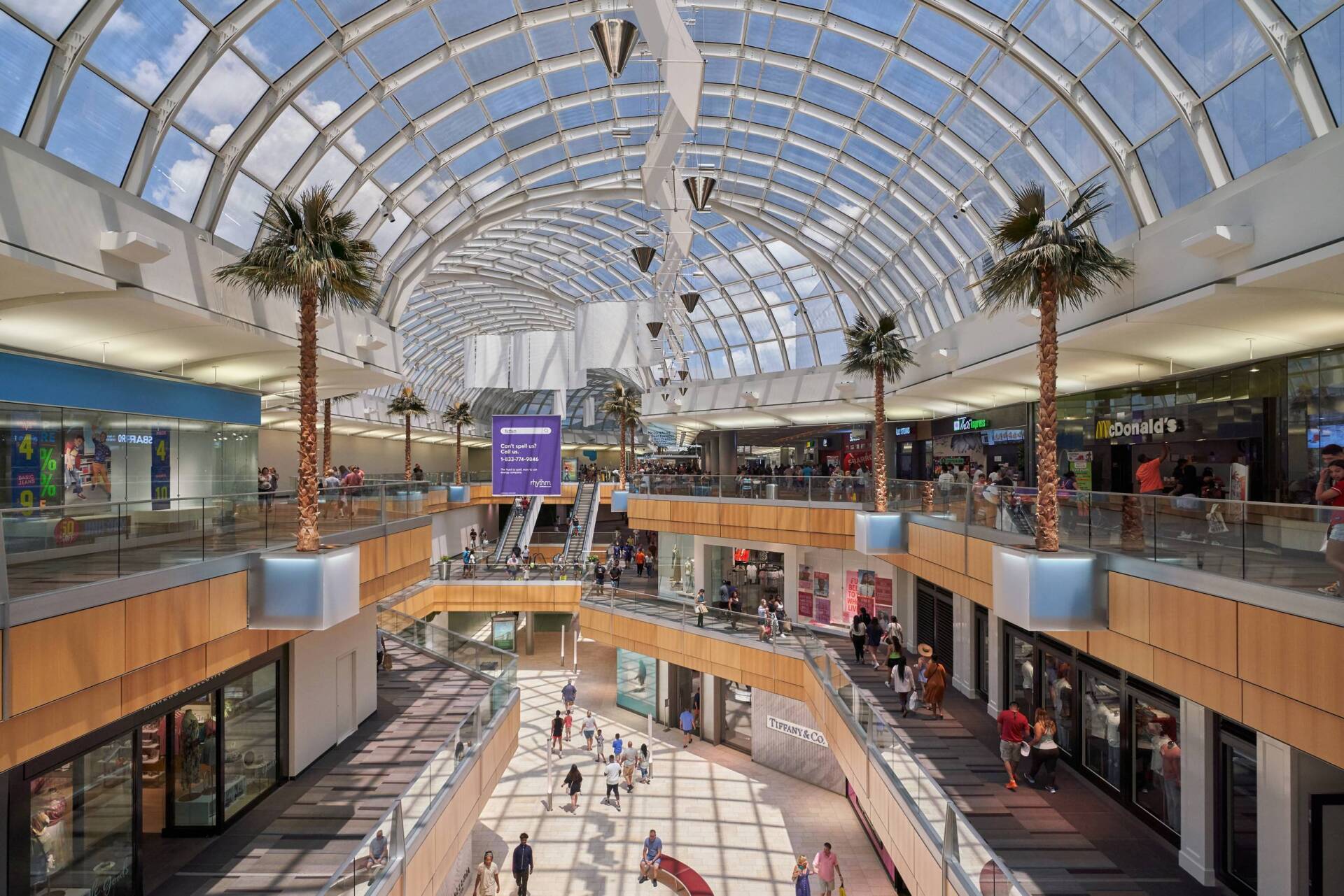 Interior view of a modern shopping mall with a glass arched roof and multiple levels of shops and walkways.