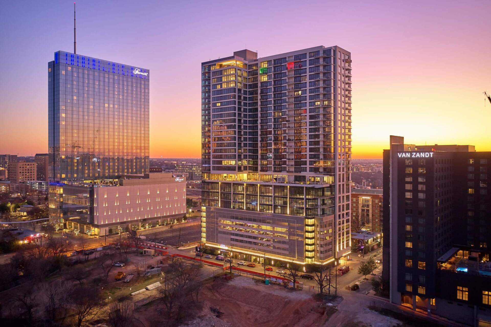 High-rise buildings at sunset, with a blue-lit tower on the left and a glass-walled building in front.