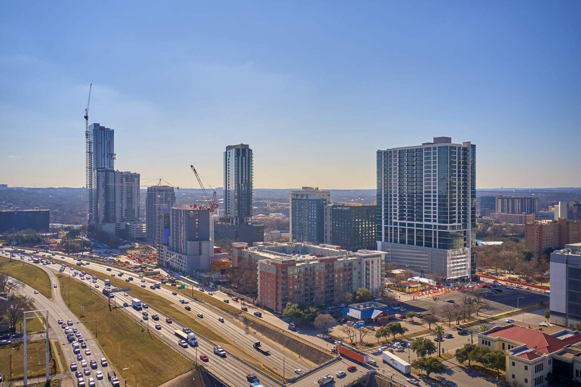 City skyline with tall buildings, construction cranes, and highway with traffic under a blue sky.