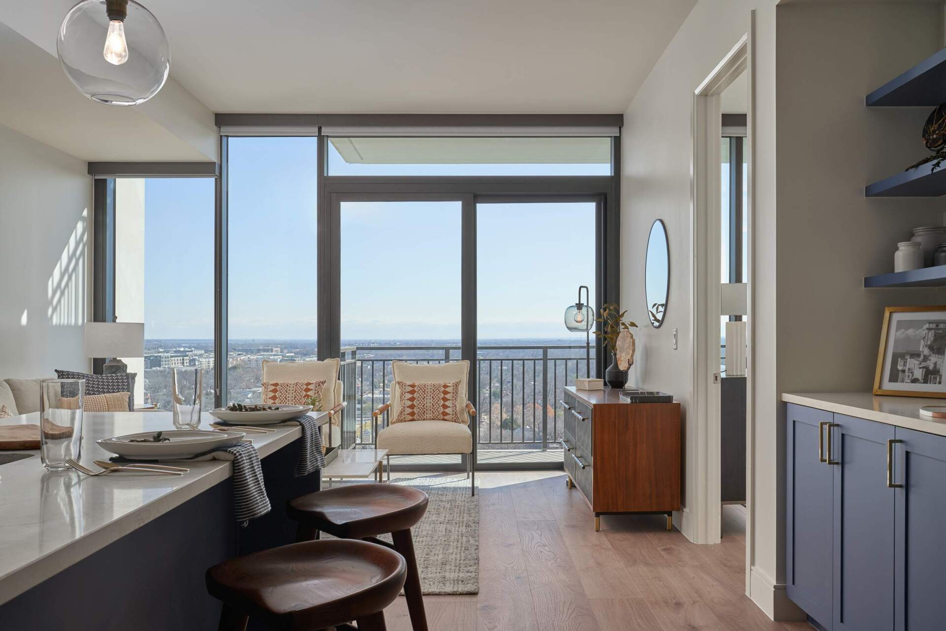 Modern apartment interior with city view through large windows; bar stools, chairs, and cabinets present.