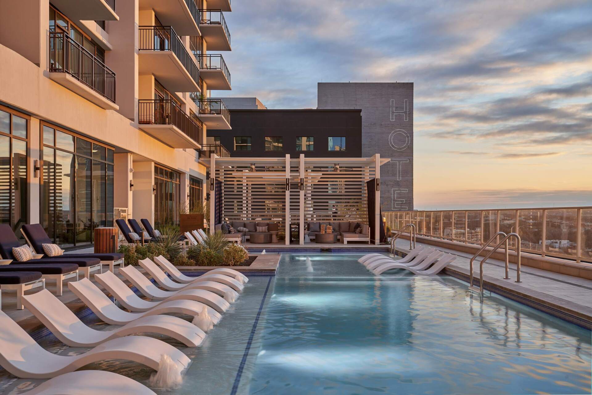 Rooftop pool with white lounge chairs, next to an apartment building and a pergola-covered seating area, with a sunset sky.