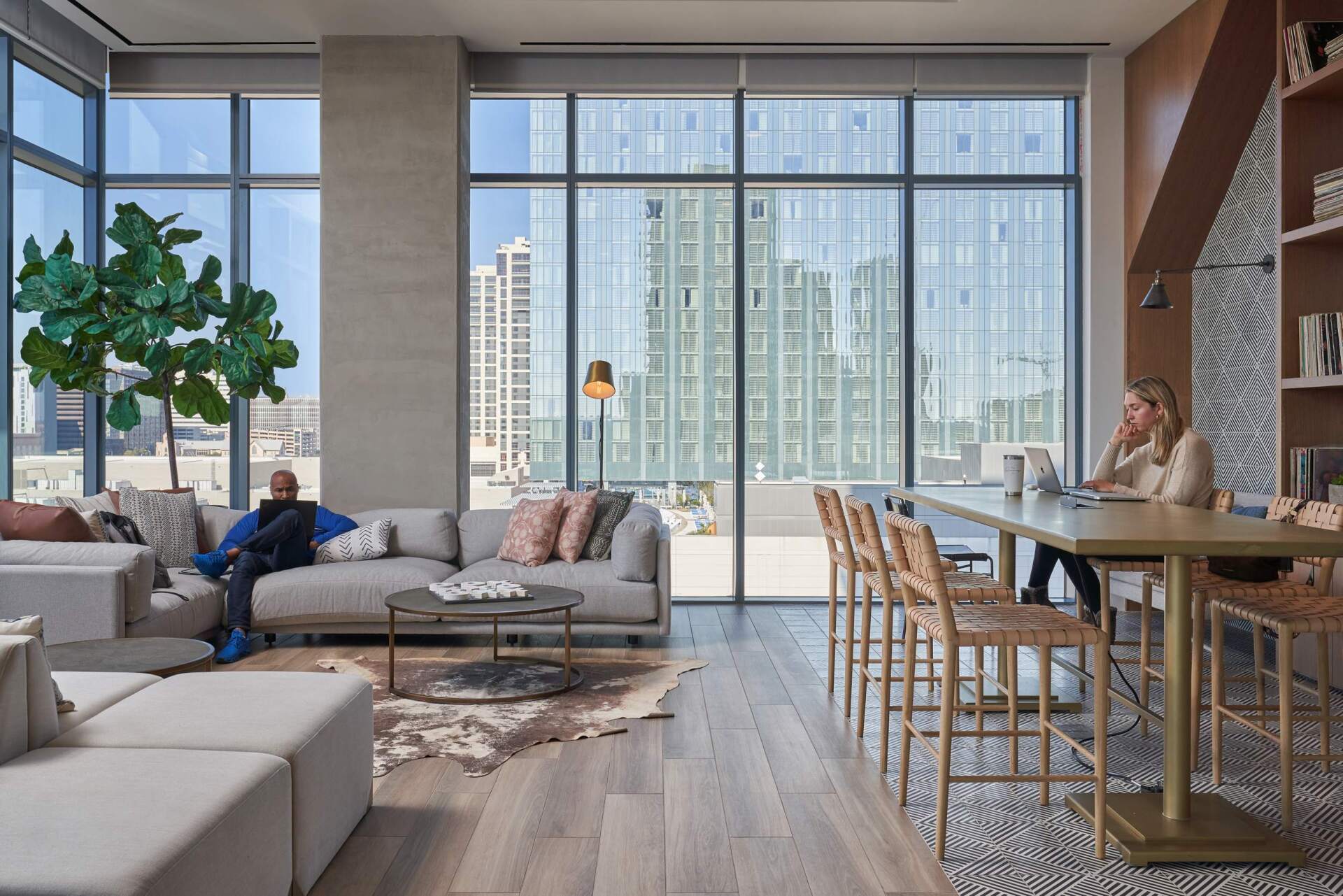 Modern office lounge with city view, sofa, table, chairs. A person sits on sofa. Another works at table.