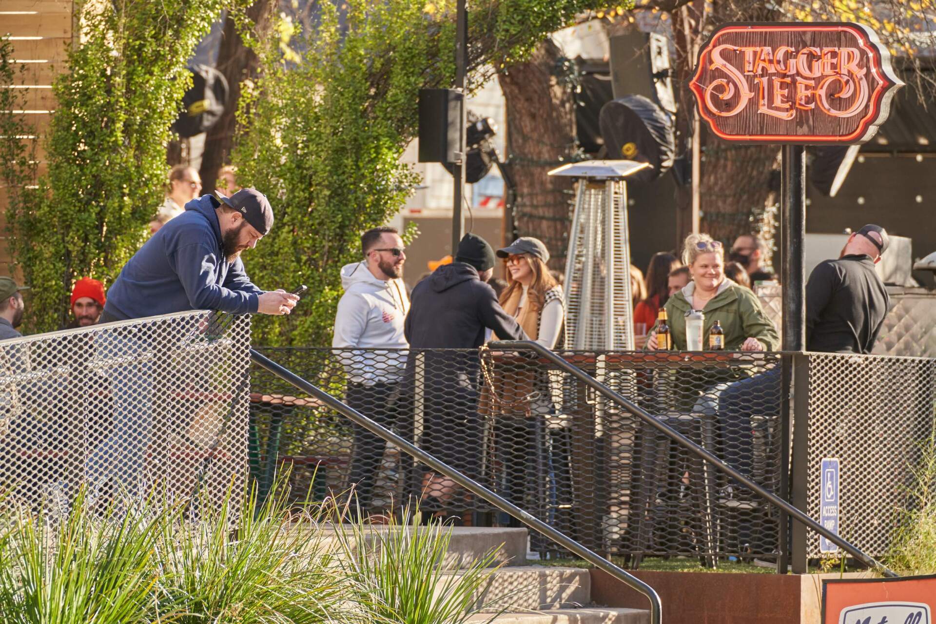 People at outdoor bar, some standing and talking at a high-top table, others looking at phones or drinking.