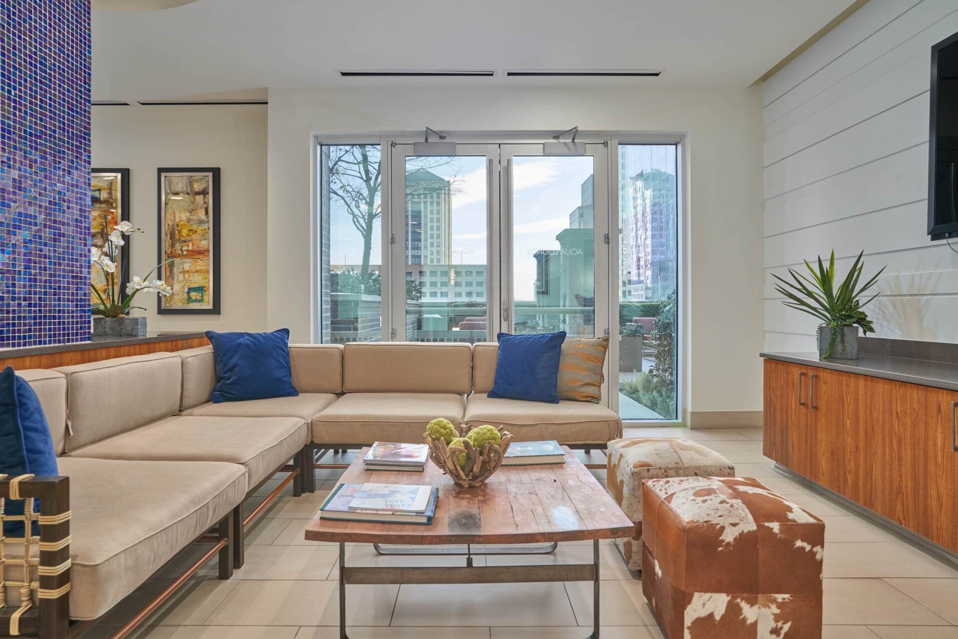 Lounge area with tan sectional sofa, blue pillows, wooden coffee table, and city view through sliding doors.