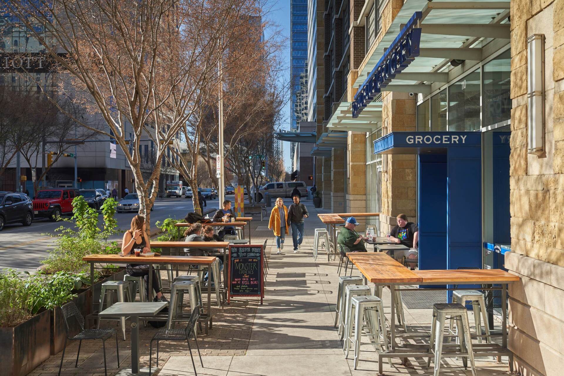 Sidewalk cafe with outdoor seating. People walking and sitting; grocery store entrance. Bright sunny day.