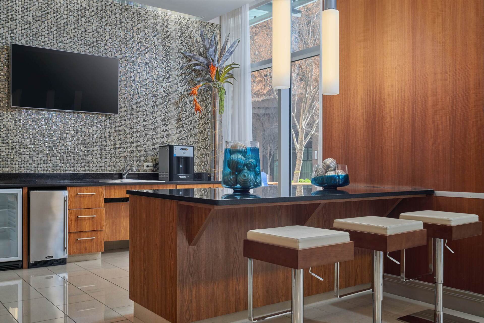 Lounge area with bar, stools, and coffee station. Features a mosaic tile wall, wood paneling, and a view of the outside.