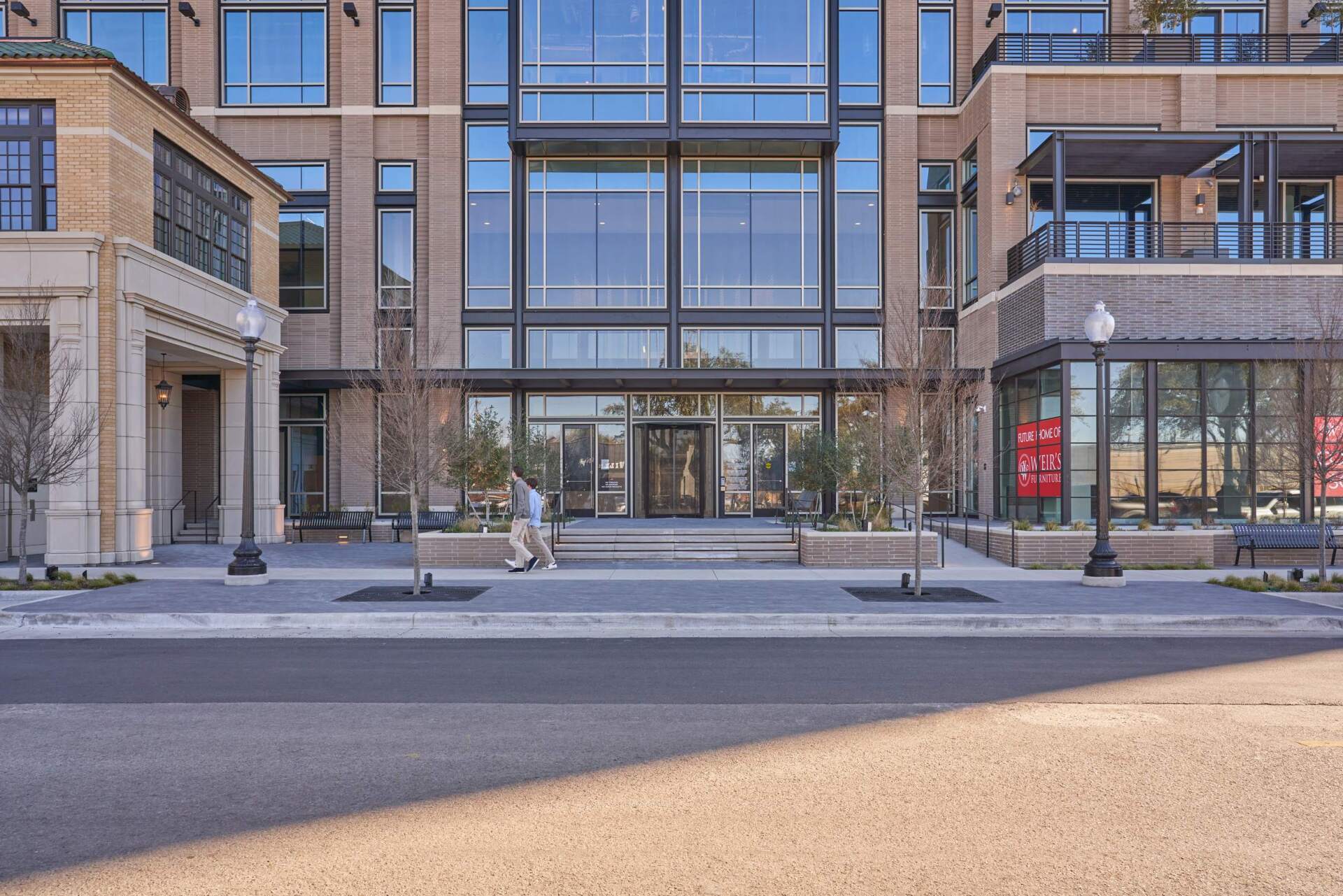 Modern building entrance with glass doors, person walking in, and small trees.