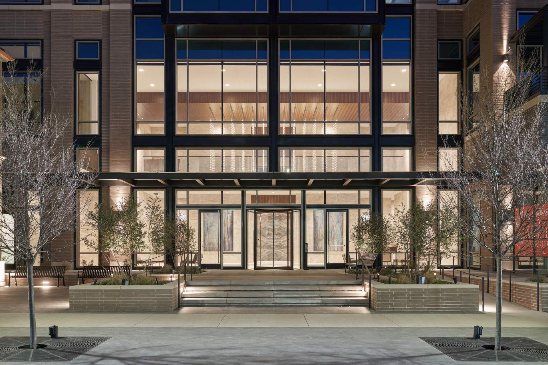 Modern building entrance with glass doors, large windows, and trees in planters.