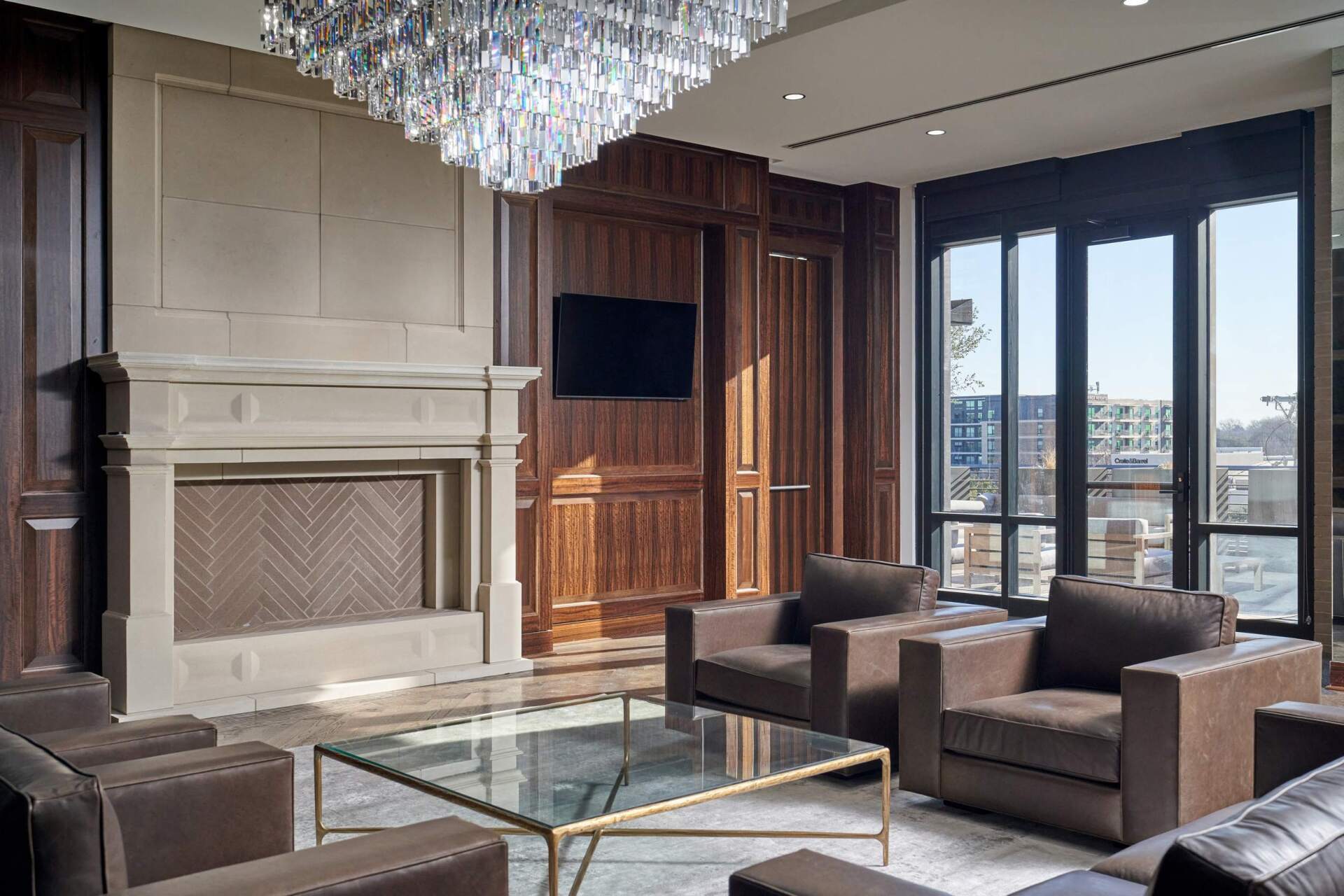 Living room with fireplace, armchairs, glass coffee table, large windows, and a chandelier.