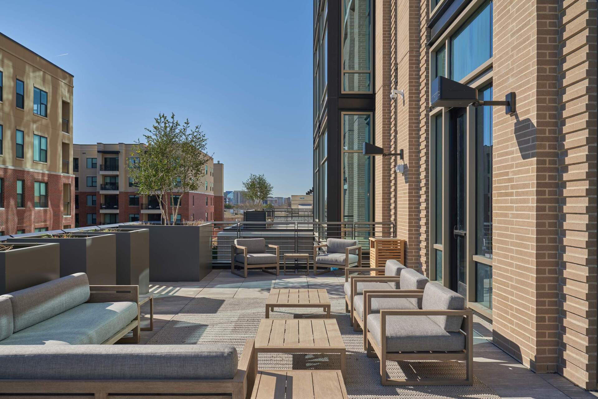 Rooftop patio with gray couches and wooden tables; buildings in the background on a sunny day.