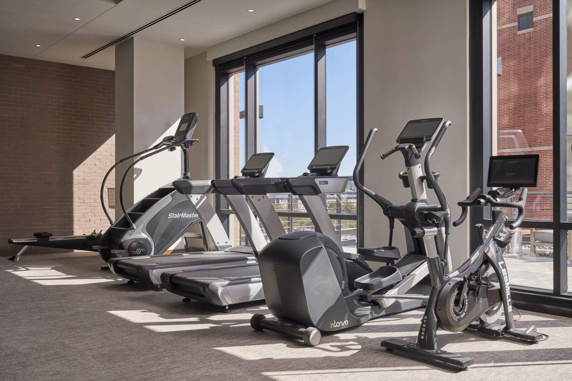 Gym with exercise equipment by a large window. Treadmills, elliptical, stationary bike.