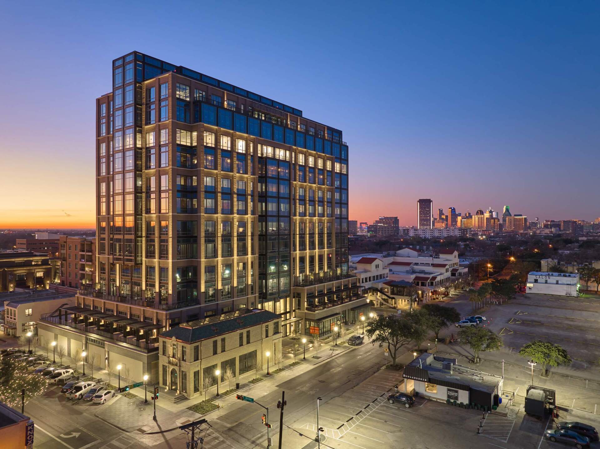 Modern high-rise building lit at dusk, with city skyline in background, street and cars below.