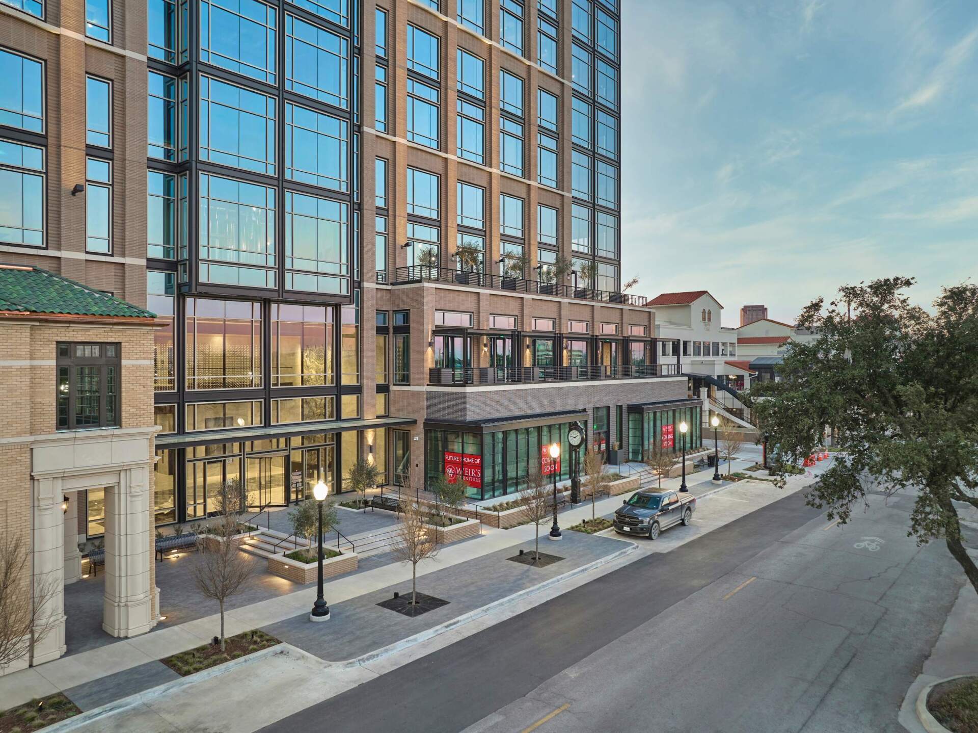 Modern building with glass facade, street view, several retail storefronts, trees, and parked vehicles.
