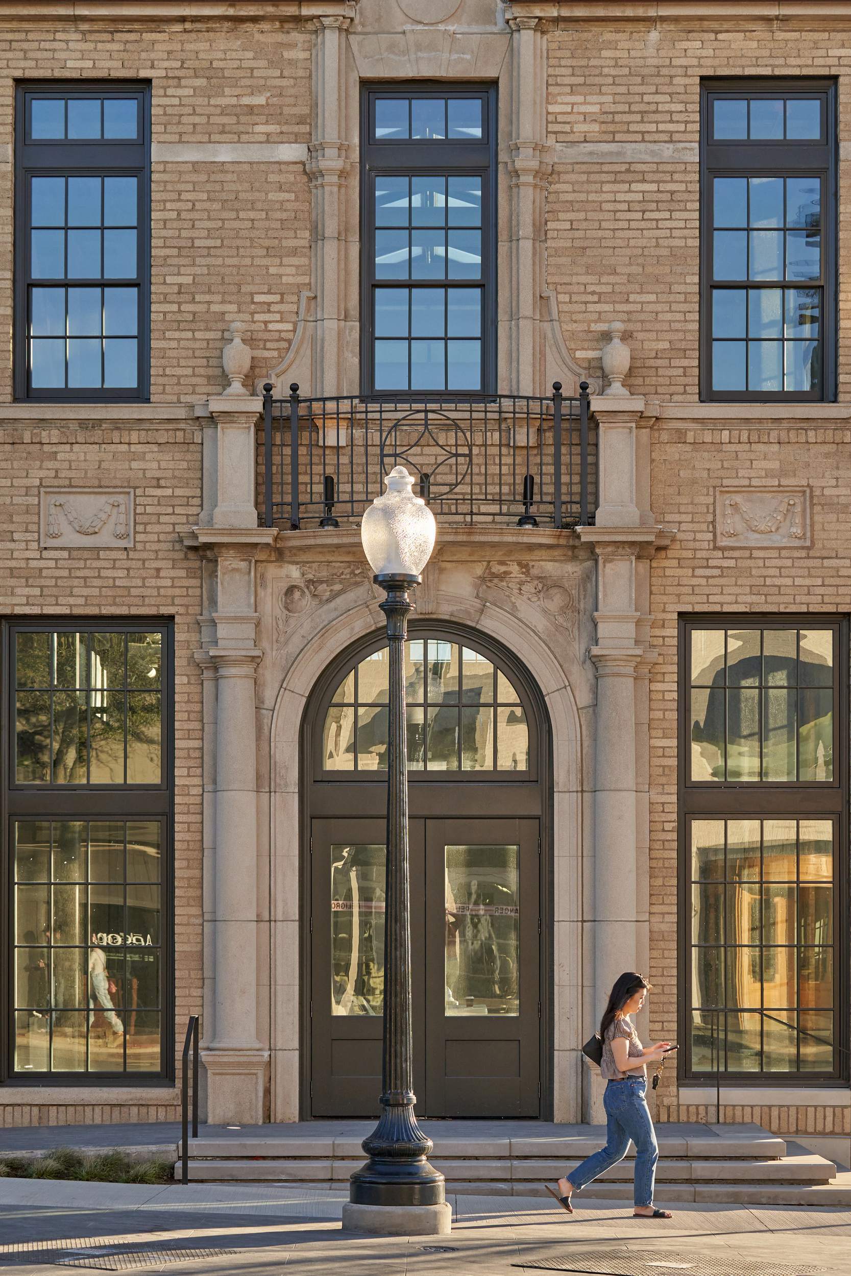 Woman walks past ornate building with tall windows and a lamppost.
