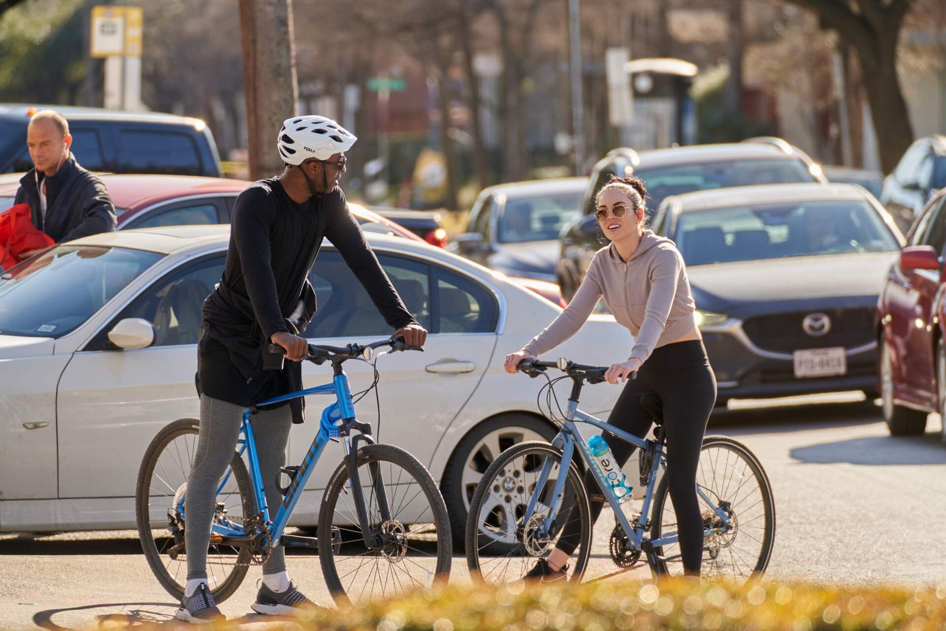 Two cyclists beside their bikes, conversing next to parked cars on a city street.