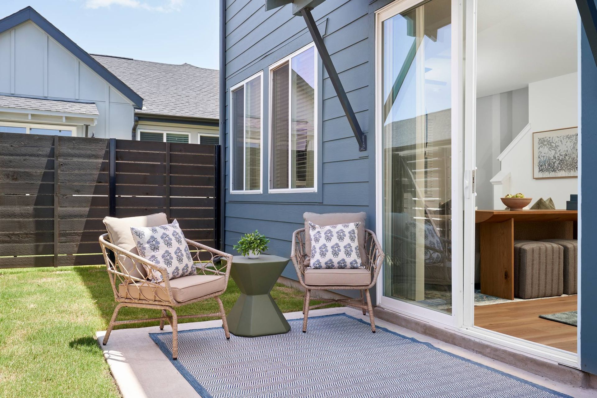 Patio with two chairs, small table, and a blue rug, next to a sliding glass door and blue house.
