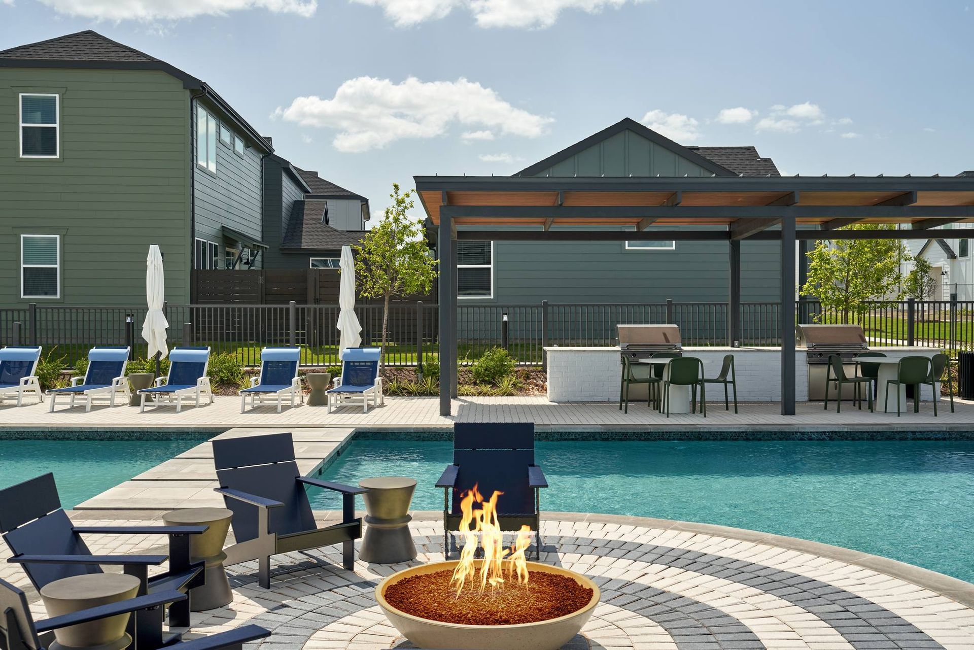 Pool area with lounge chairs, fire pit, and outdoor kitchen, in front of green buildings.