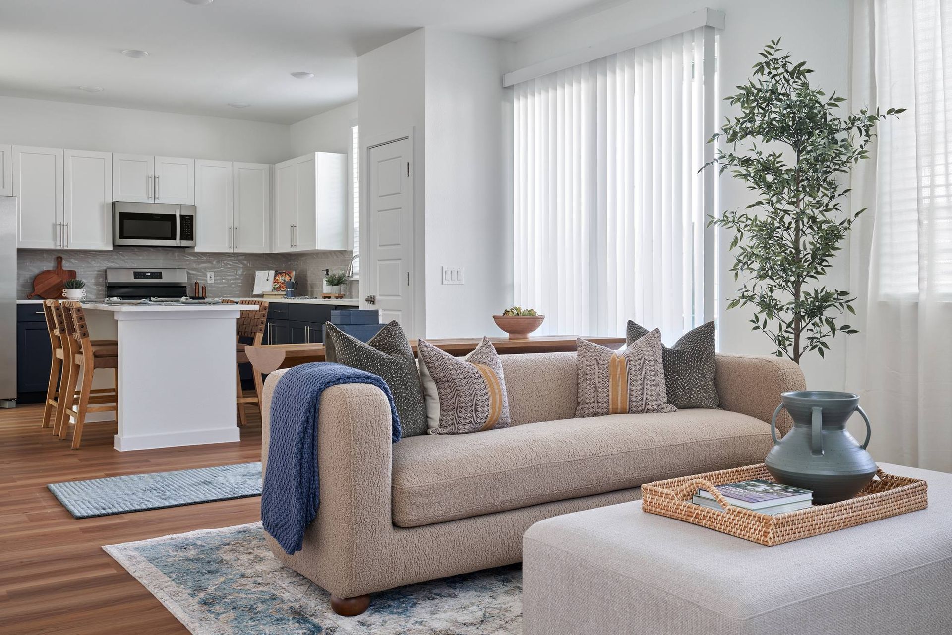 Living room with a kitchen in the background. Beige sofa, ottoman, and hardwood floors.