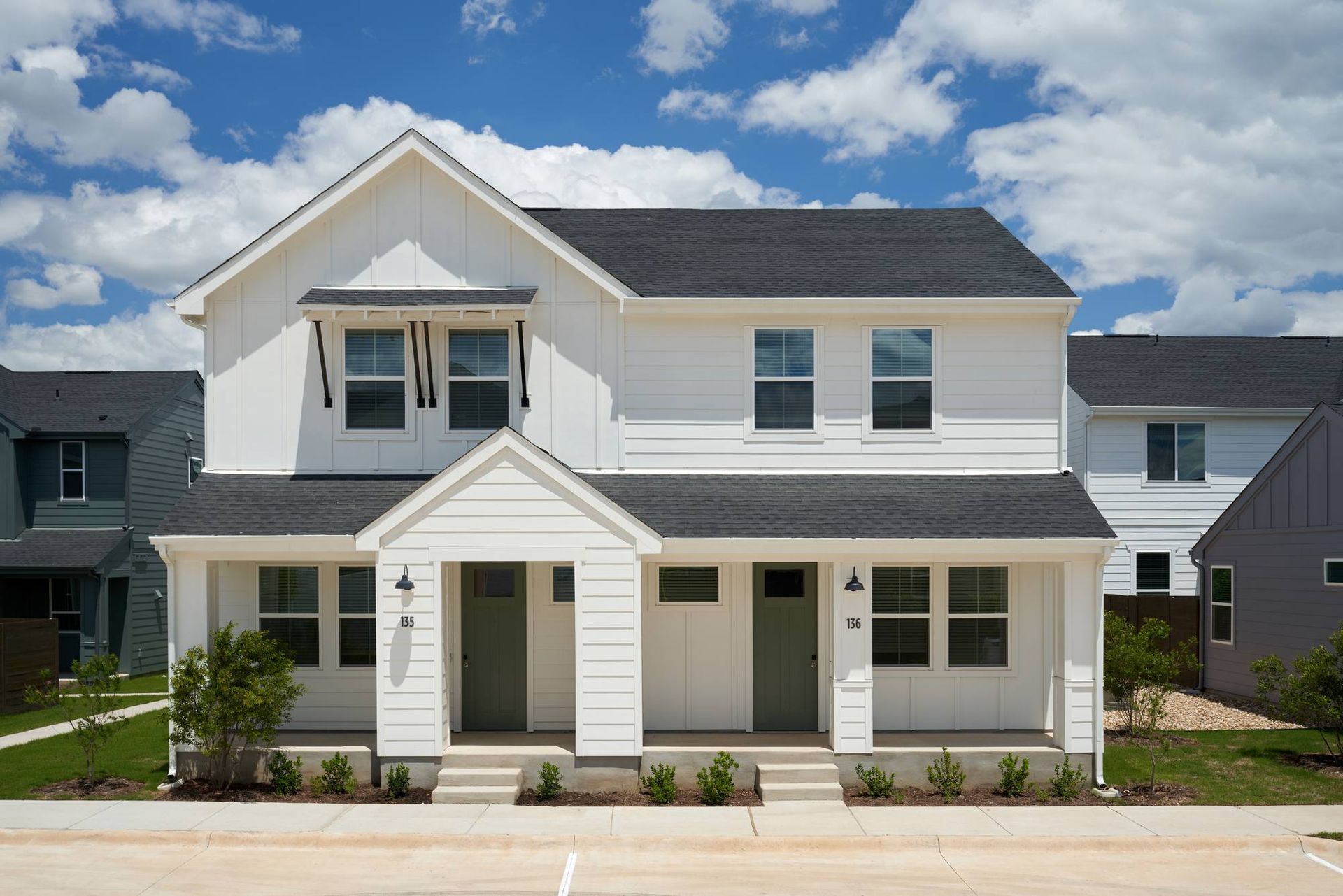 White two-story duplex with dark roof and green doors against a blue sky.