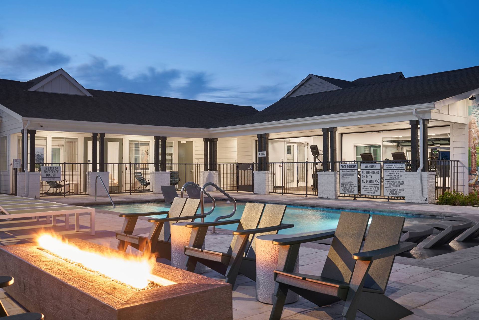 A pool area at dusk with a lit fire pit and Adirondack chairs facing the water. Building in background.
