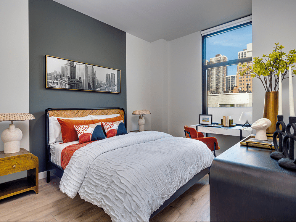 Apartment bedroom with bed, wall art, night stand, table, chair, oversized window, and dresser at Gild Apartment.